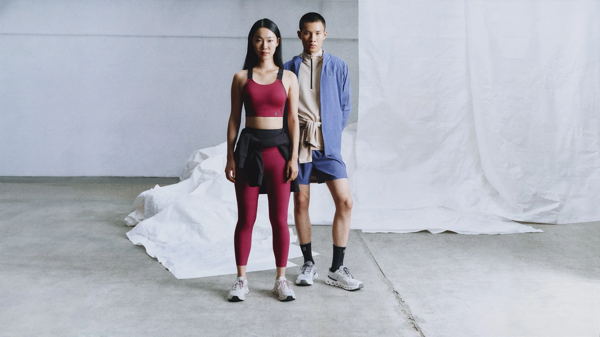 Zwei Personen in Sportbekleidung stehen vor einem weißen Hintergrund. Die Frau trägt rote Leggings und einen Sport-BH, der Mann Shorts und ein Oberteil.